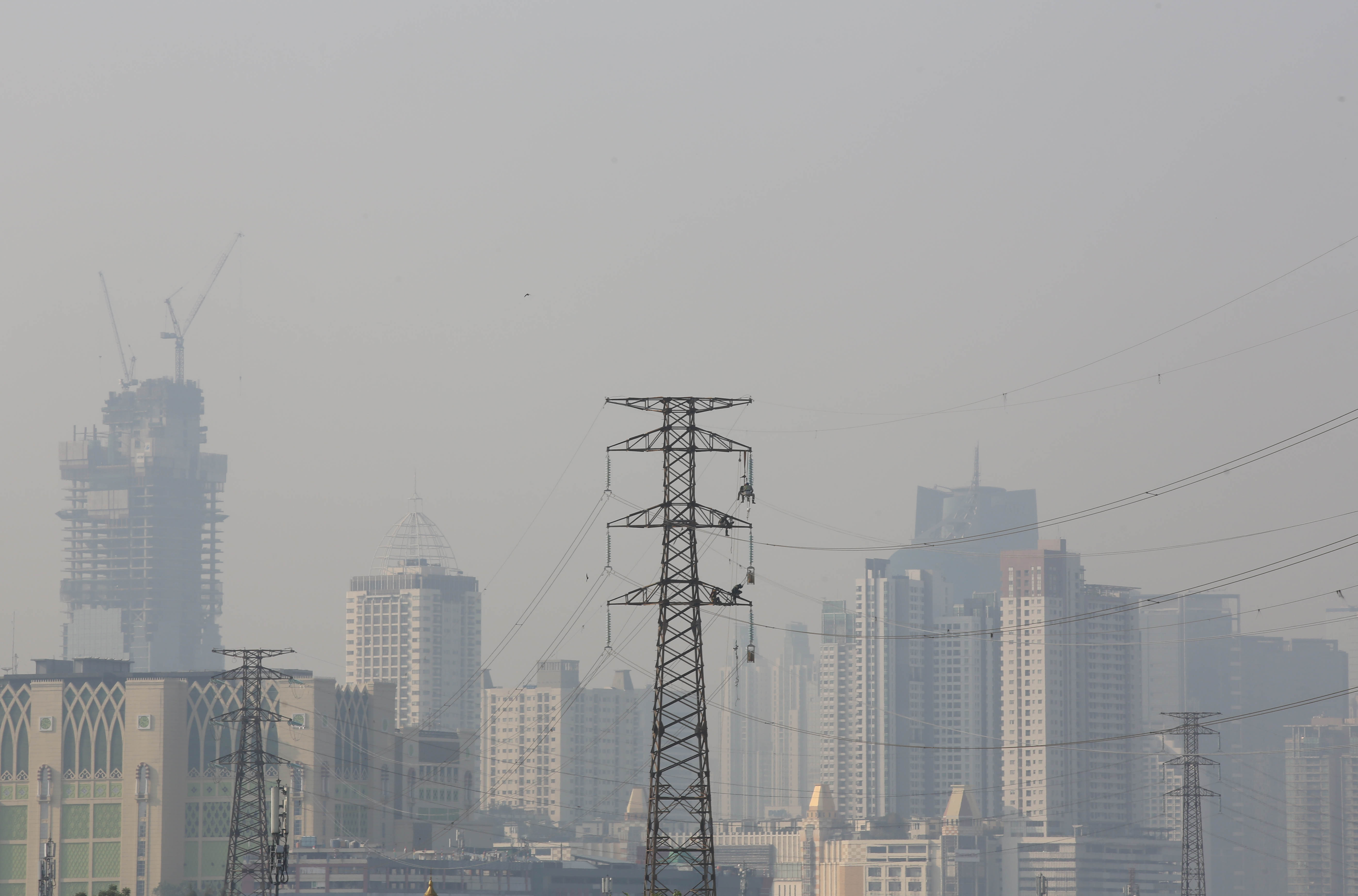 Pekerja melakukan pemeliharaan aliran SUTET dengan latar belakang polusi udara di Jakarta