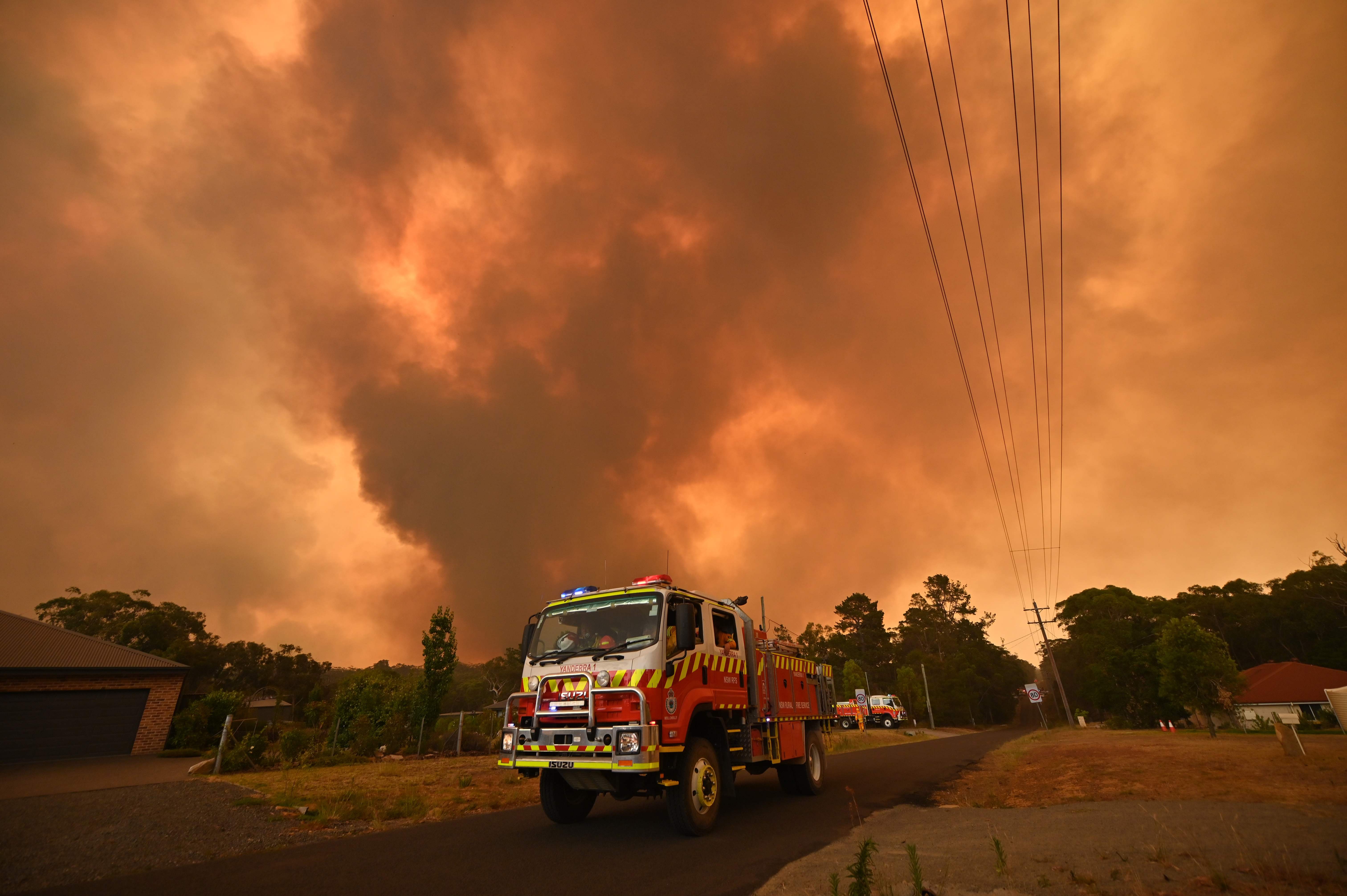 Truk pemadam kebakaran bersiaga di sebuah jalan dan tampak kepulan kebakaran semak-semak di Bargo, Sydney, Australia.