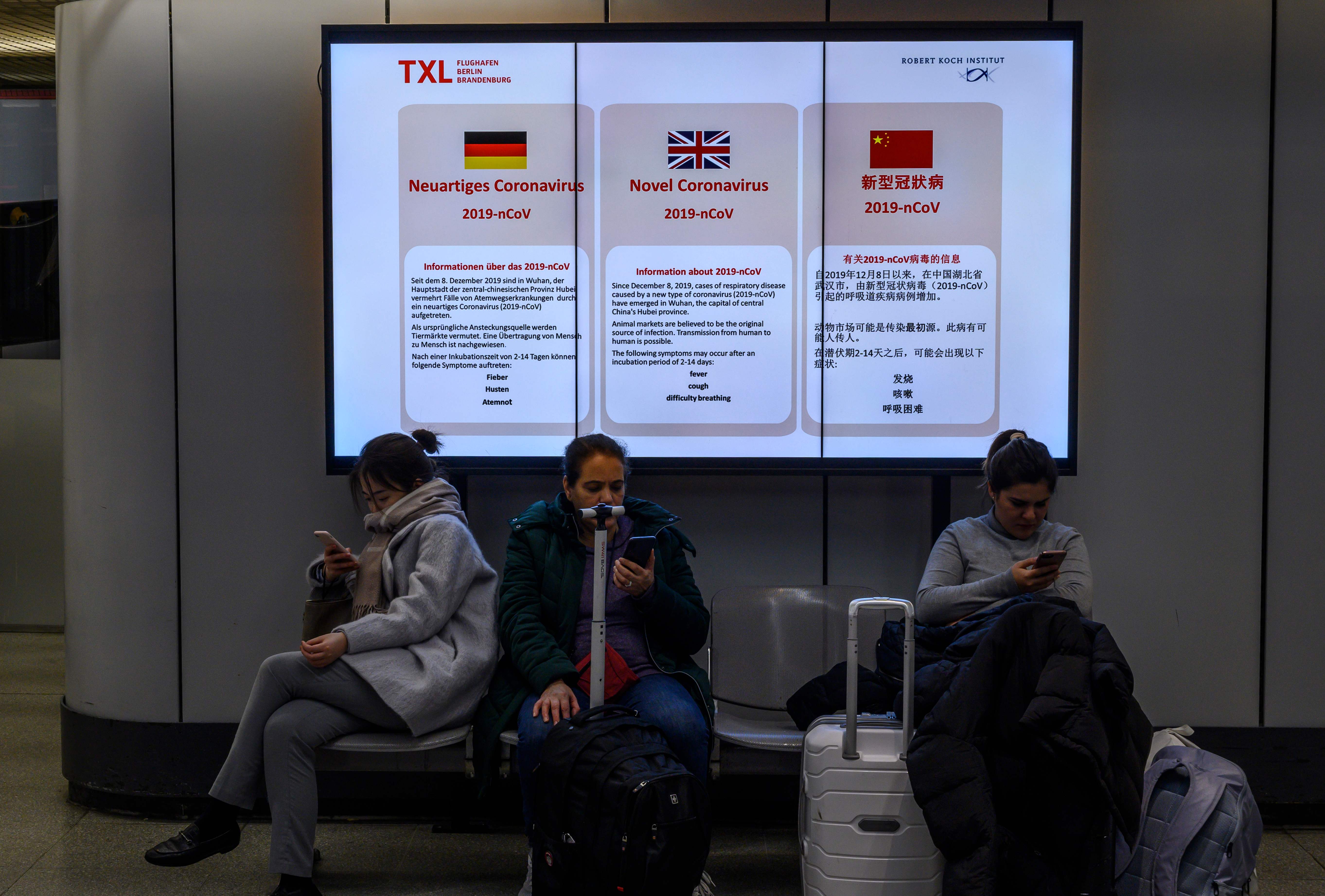 Penumpang sedang duduk di depan billboard yang menginformasikan virus korona di bandara Berlin