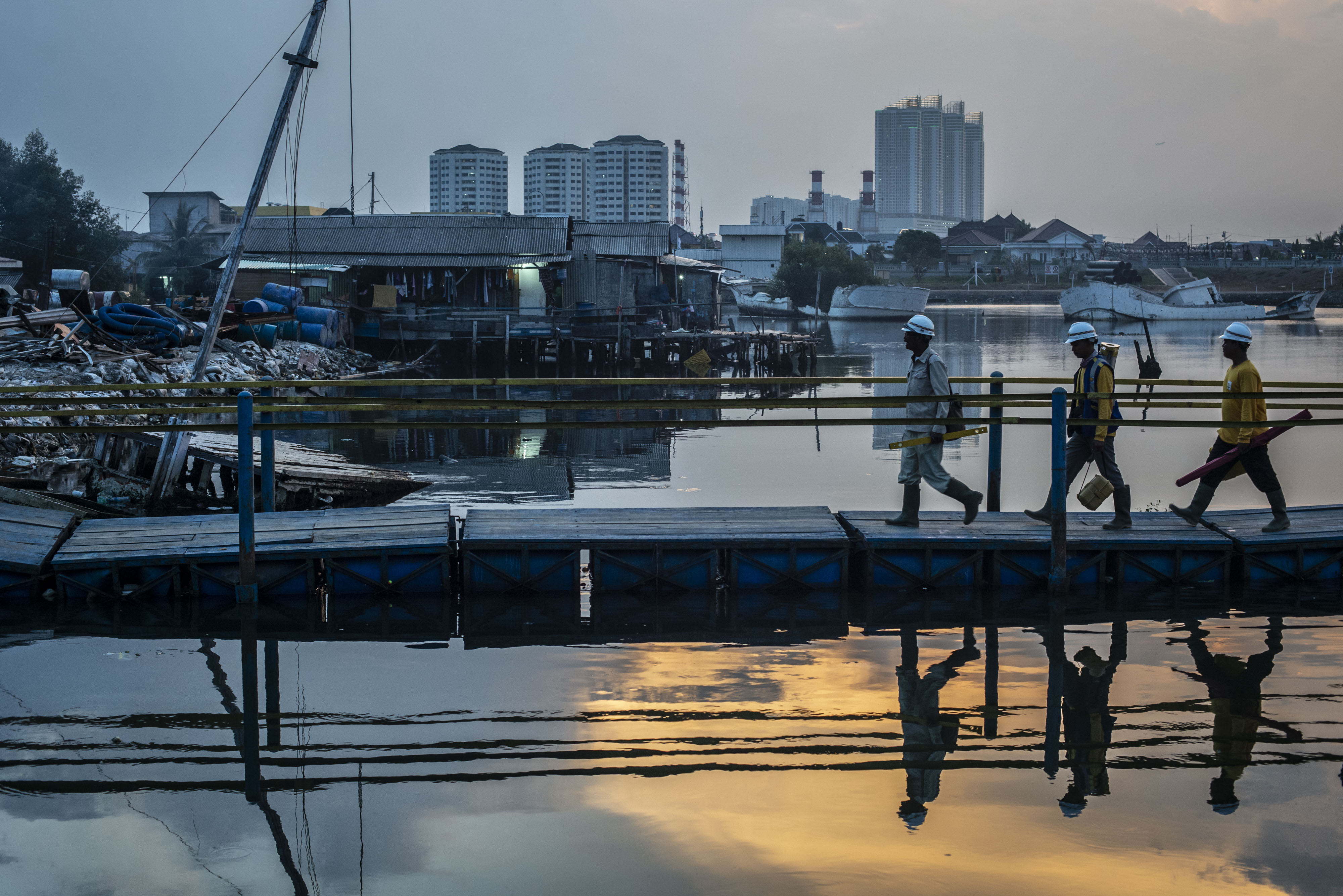 Sejumlah warga berjalan di jembatan dengan latar belakang permukiman semi permanen di kawasan pesisir Muara Baru, Jakarta.