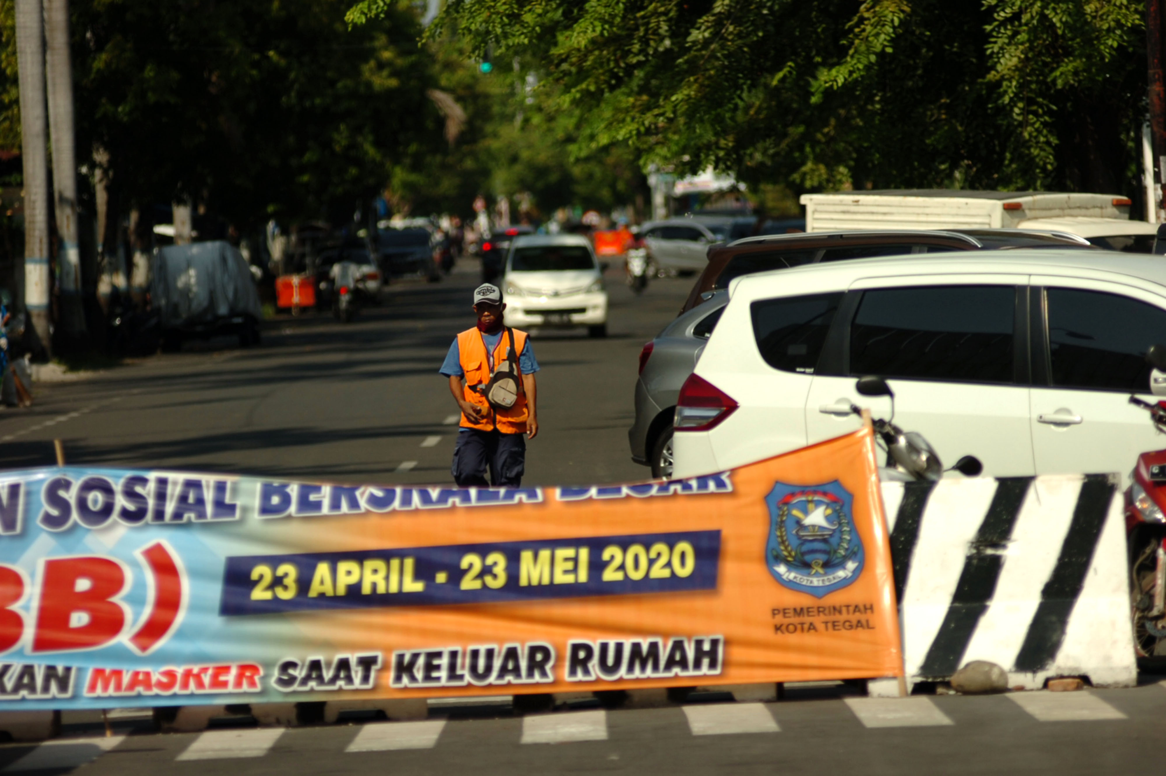 Seorang juru parkir berada samping spanduk Pembatasan Sosial Berskala Besar  di beton Jalan Hos Cokroaminoto, Tegal,J Jawa Tengah.