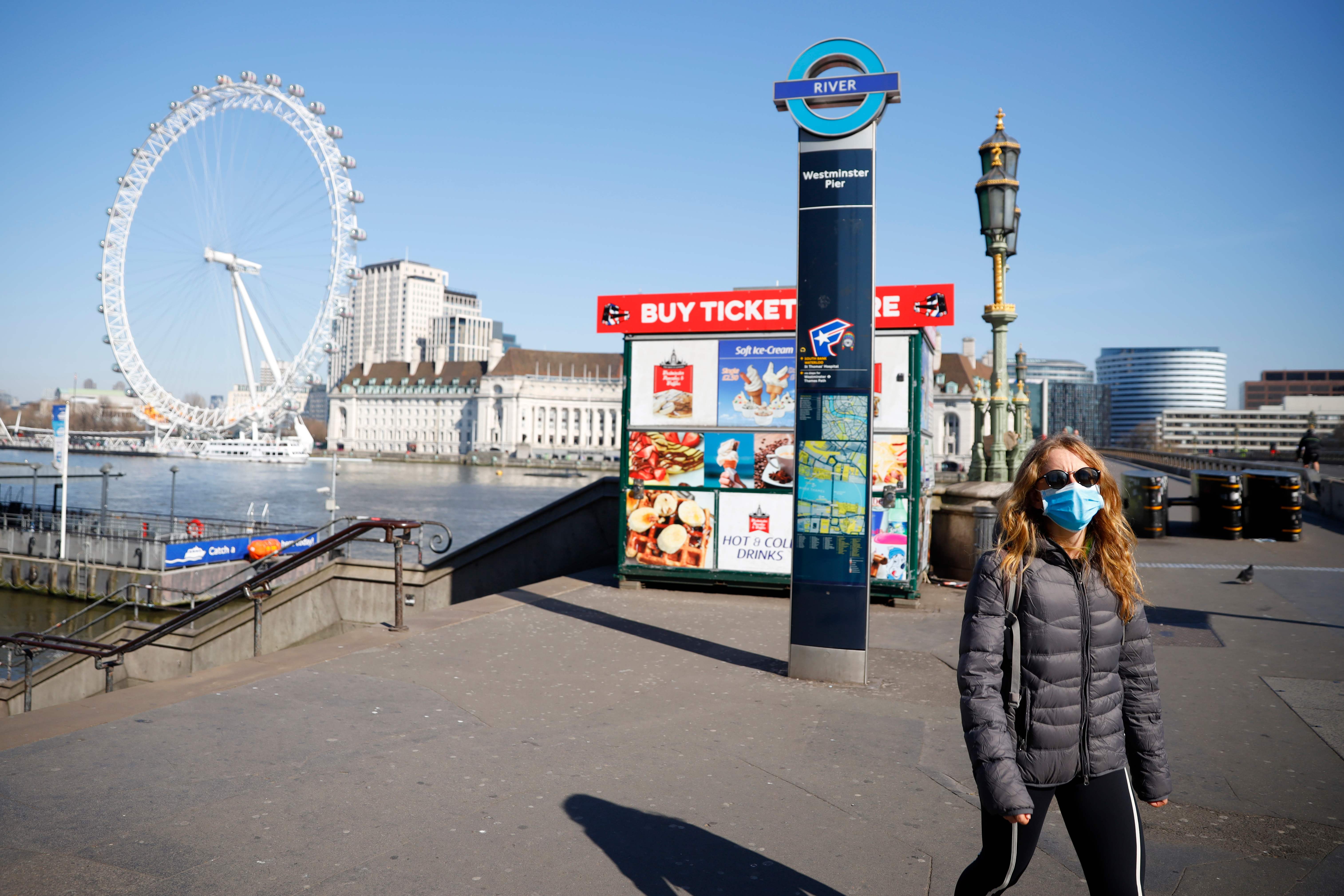 Dengan mengenakan masker, seorang warga berjalan di Westminster Pier yang dilatarbelakangi London Eye.