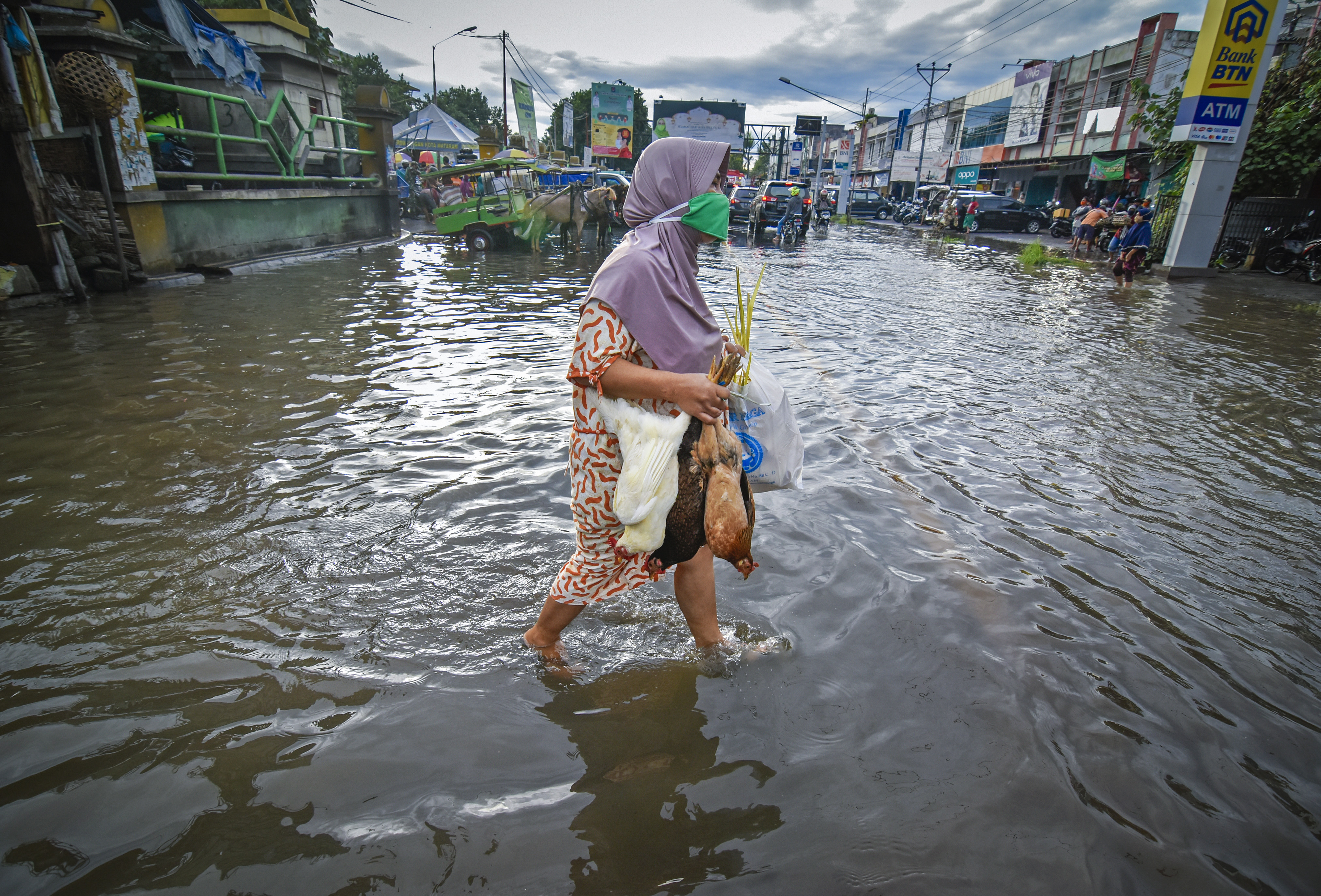 Dengan membawa ayam, seorang ibu berjalan melintasi genangan di jalan wilayah Mataram, NTB.