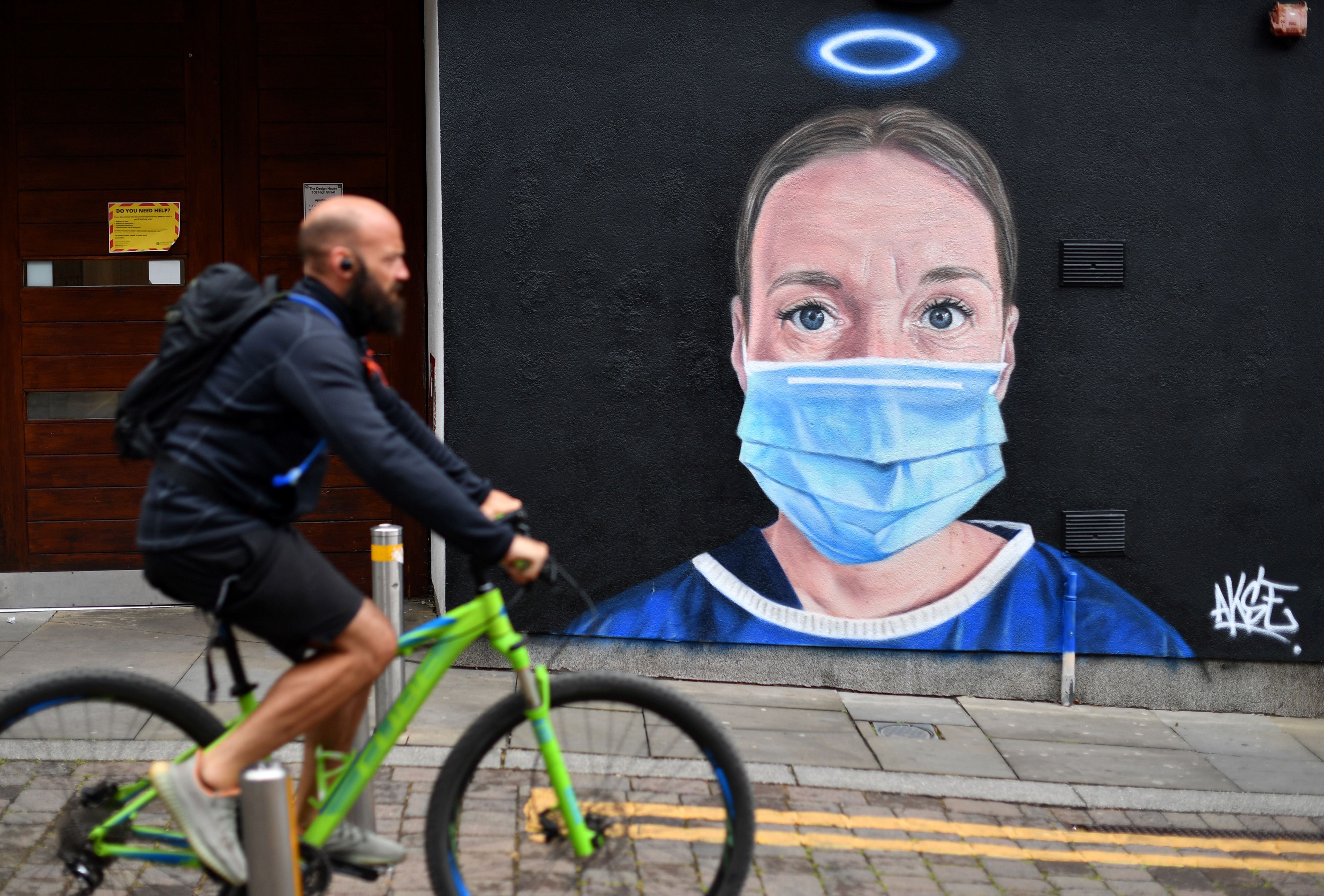 Seorang pesepeda melintasi mural tenaga medis di Manchester, Inggris.
