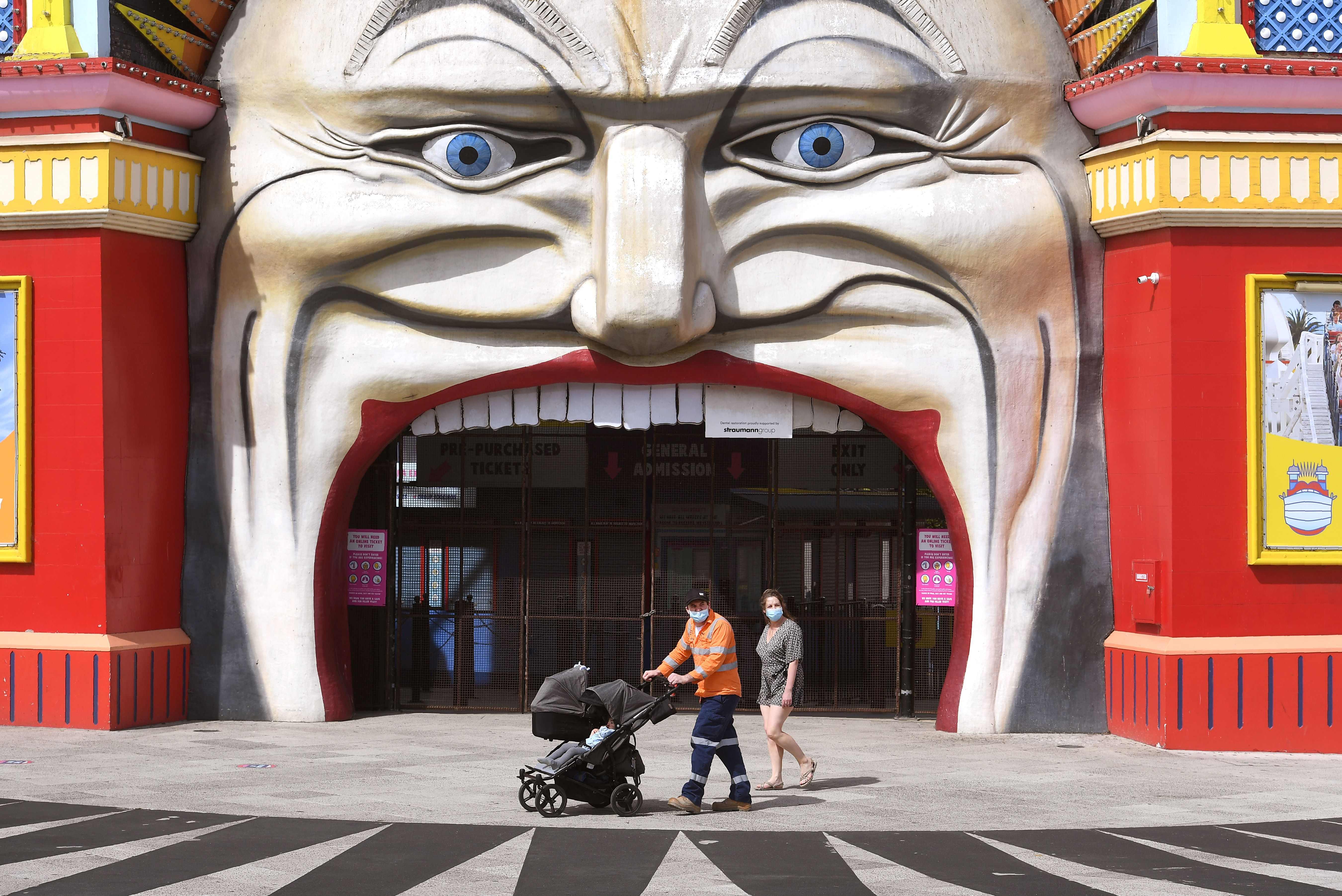 Warga mengenakan masker melintas di depan pintu masuk Luna Park, Melbourne, Australia.