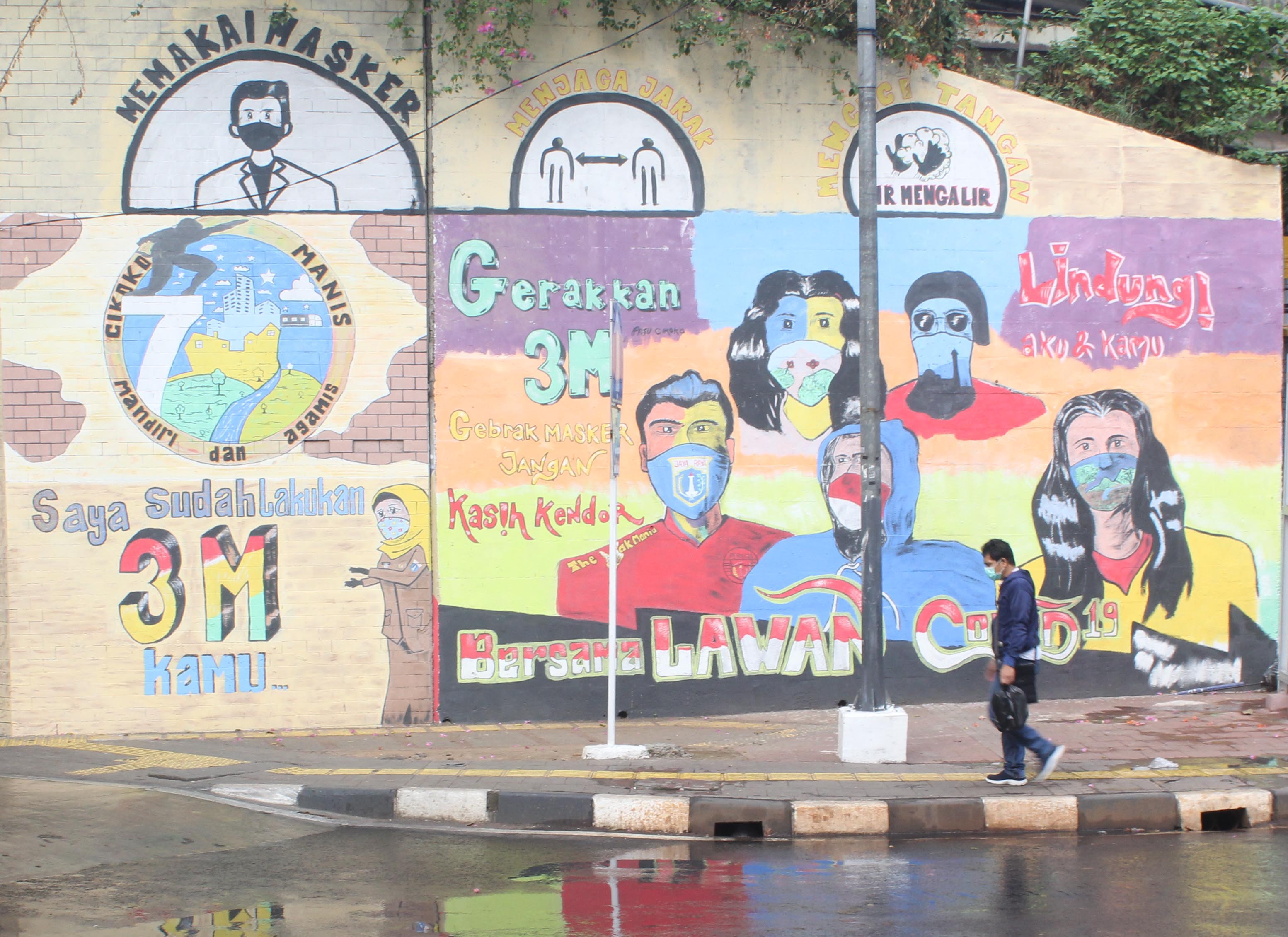 Pejalan kaki melintasi mural terkait 3M di kawasan Cawang, Jakarta.