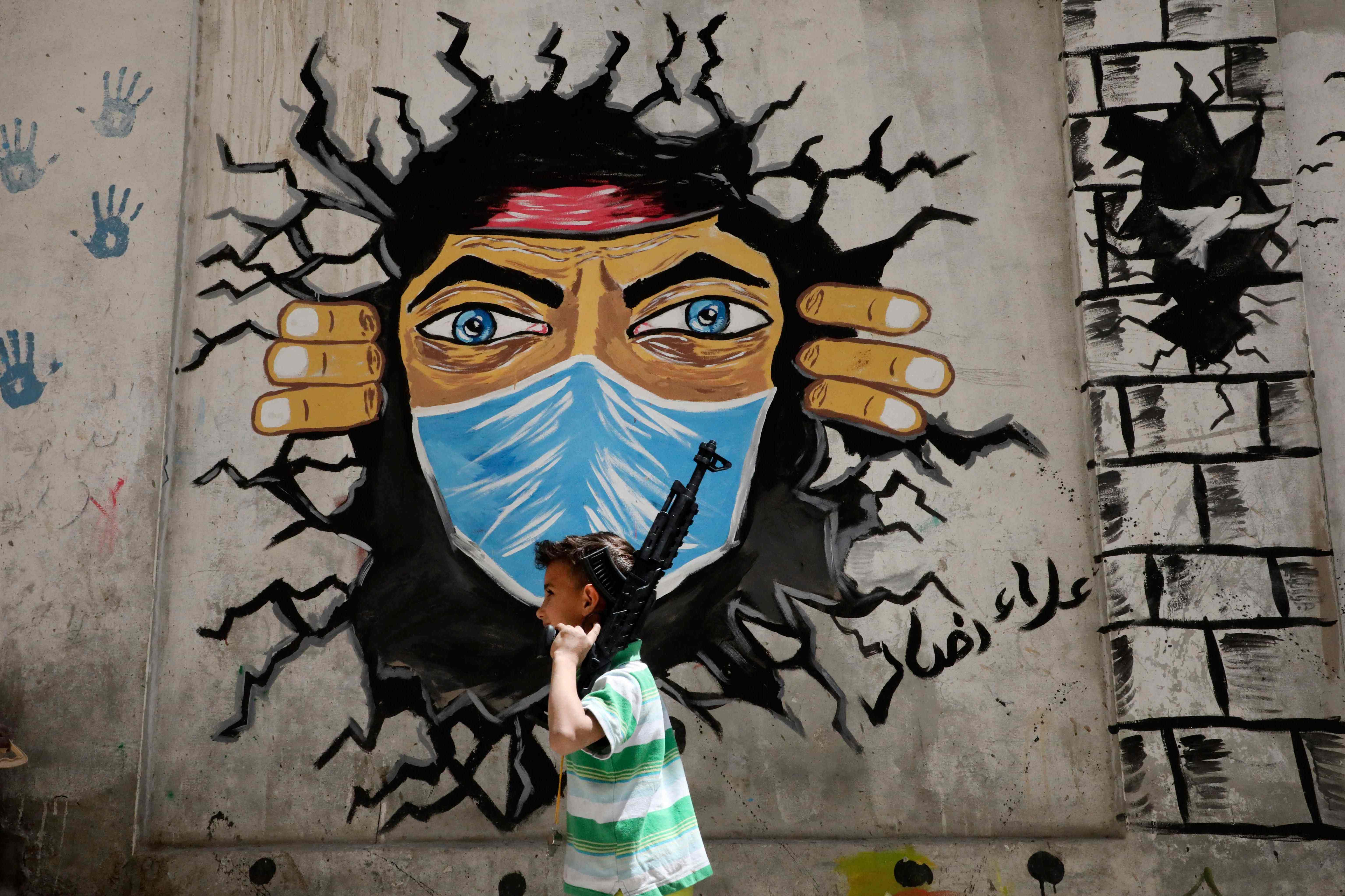 Seorang anak laki-laki Palestina berjalan dengan senapan mainan melewati mural covid-19 di kamp pengungsi al-Fawwar.