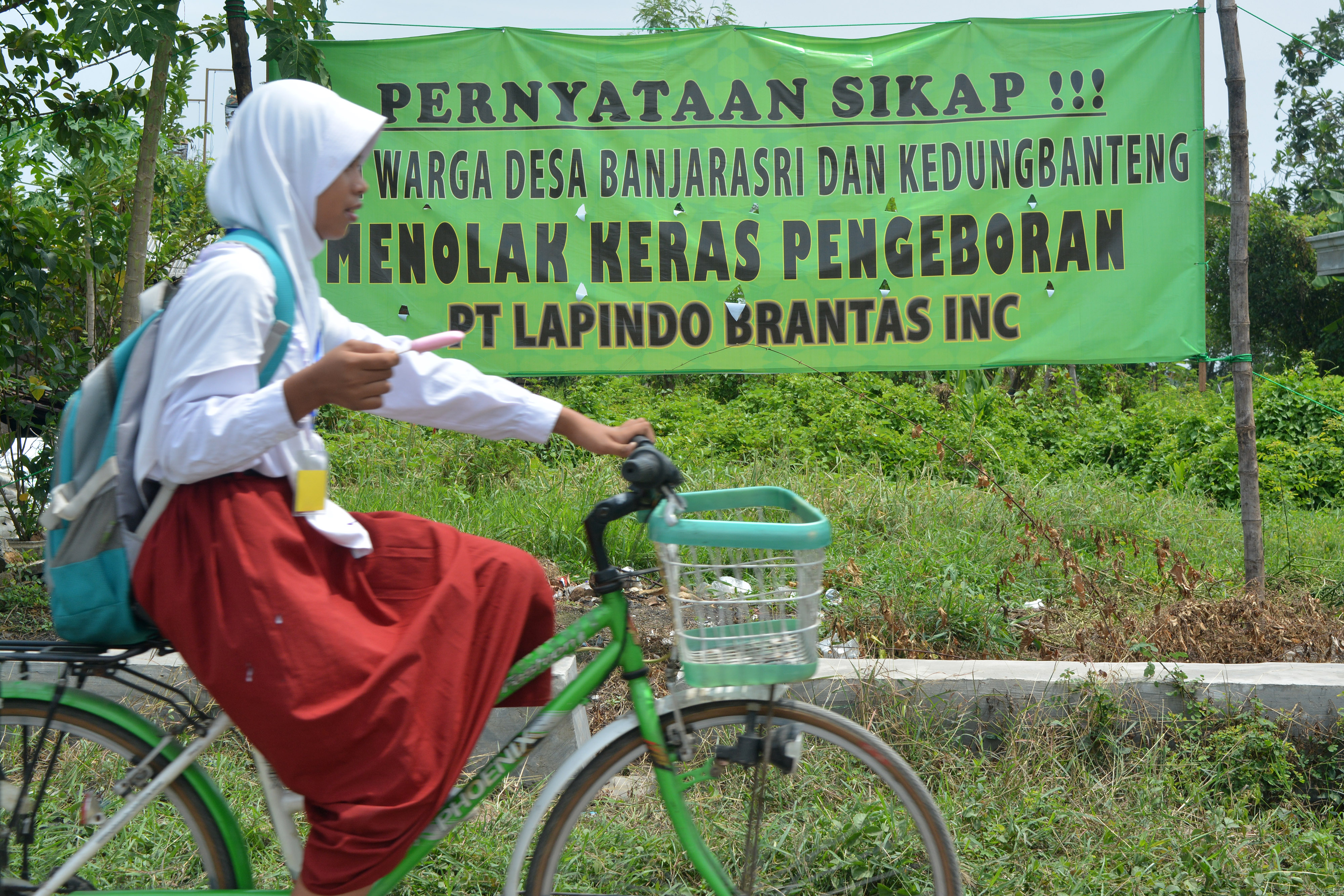 Pelajar melintas di depan spanduk penolakan pengeboran sumur gas baru Lapindo yang terpasang di Sumur Tanggulangin 1, Sidoarjo, 2019.