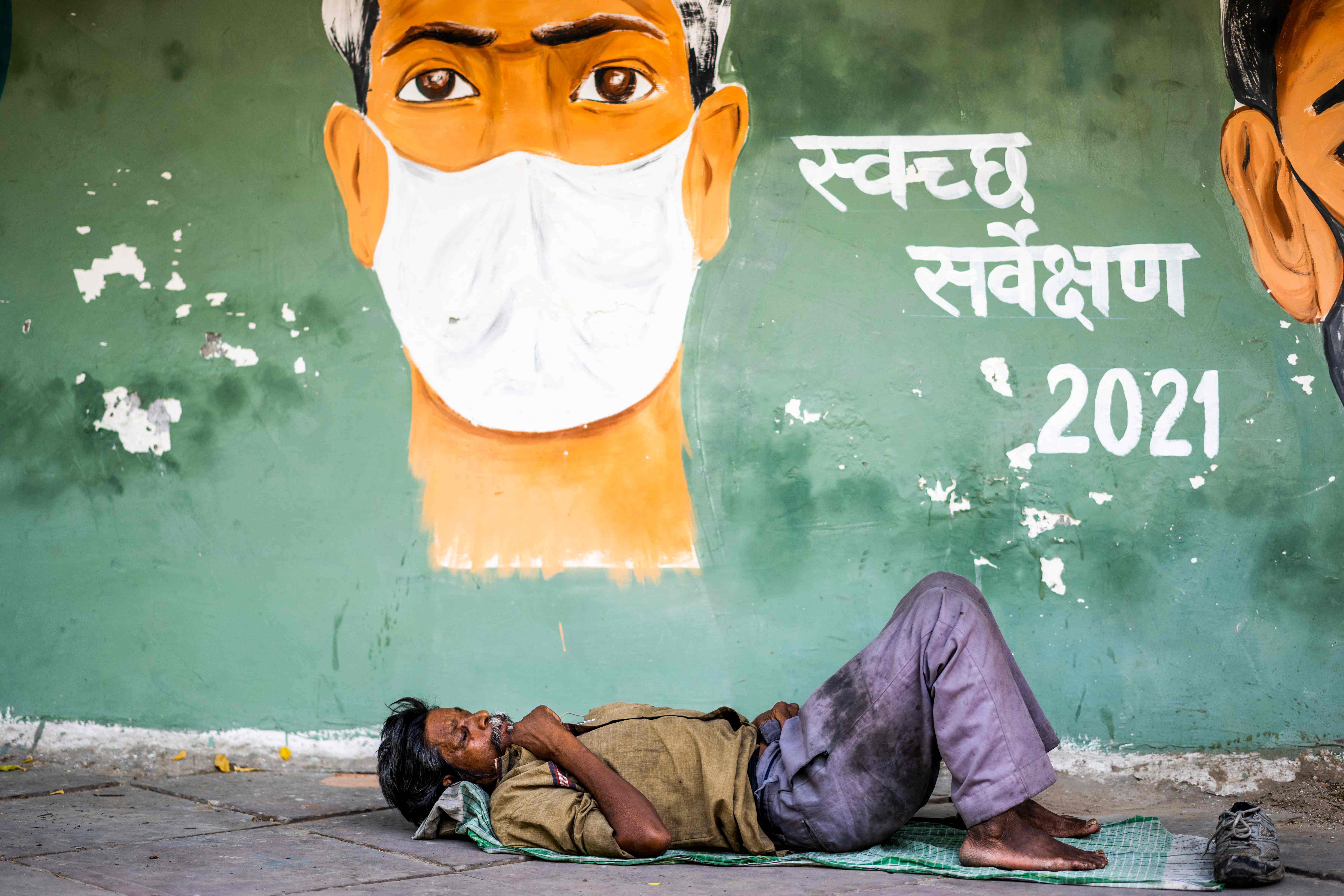 Seorang pria beristirahat di jalan setapak di bawah lukisan kesadaran bermasker di New Delhi, Selasa (11/5).