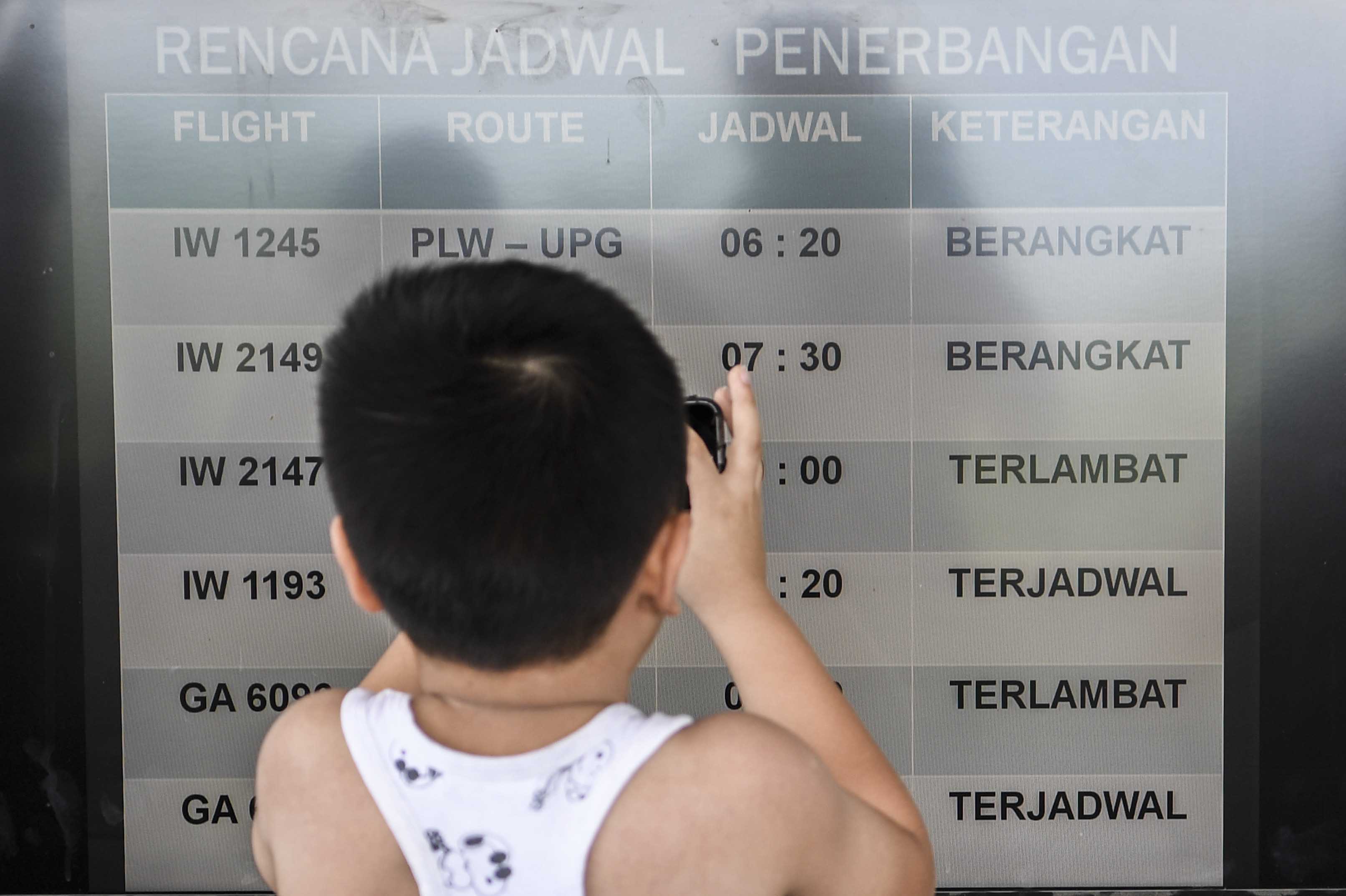 Dengan menggunakan gawai, seorang anak memotret jadwal penerbangan di bandara.