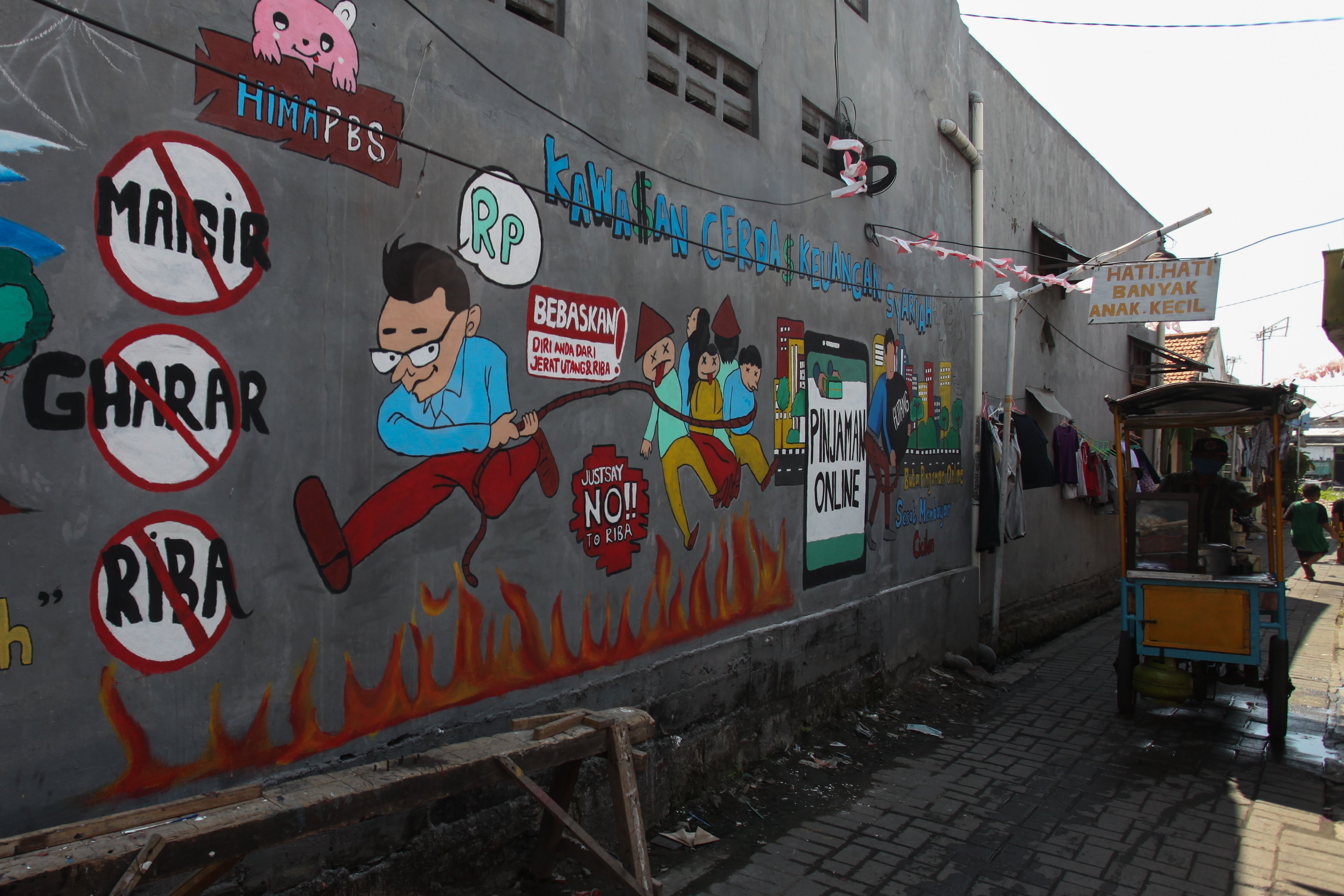 Pedagang makanan melintasi mural terkait bahaya pinjol ilegal di wilayah Surabaya, Jawa Timur.