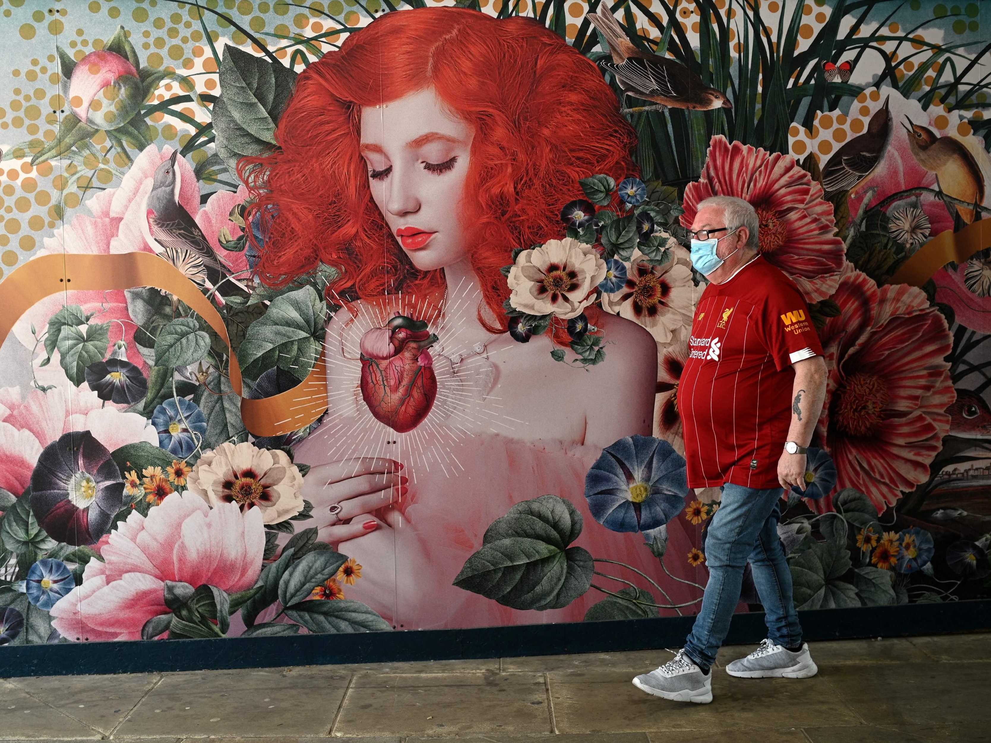 Seorang pejalan kaki yang mengenakan masker berjalan melewati mural di dinding, Blackburn, barat laut Inggris.