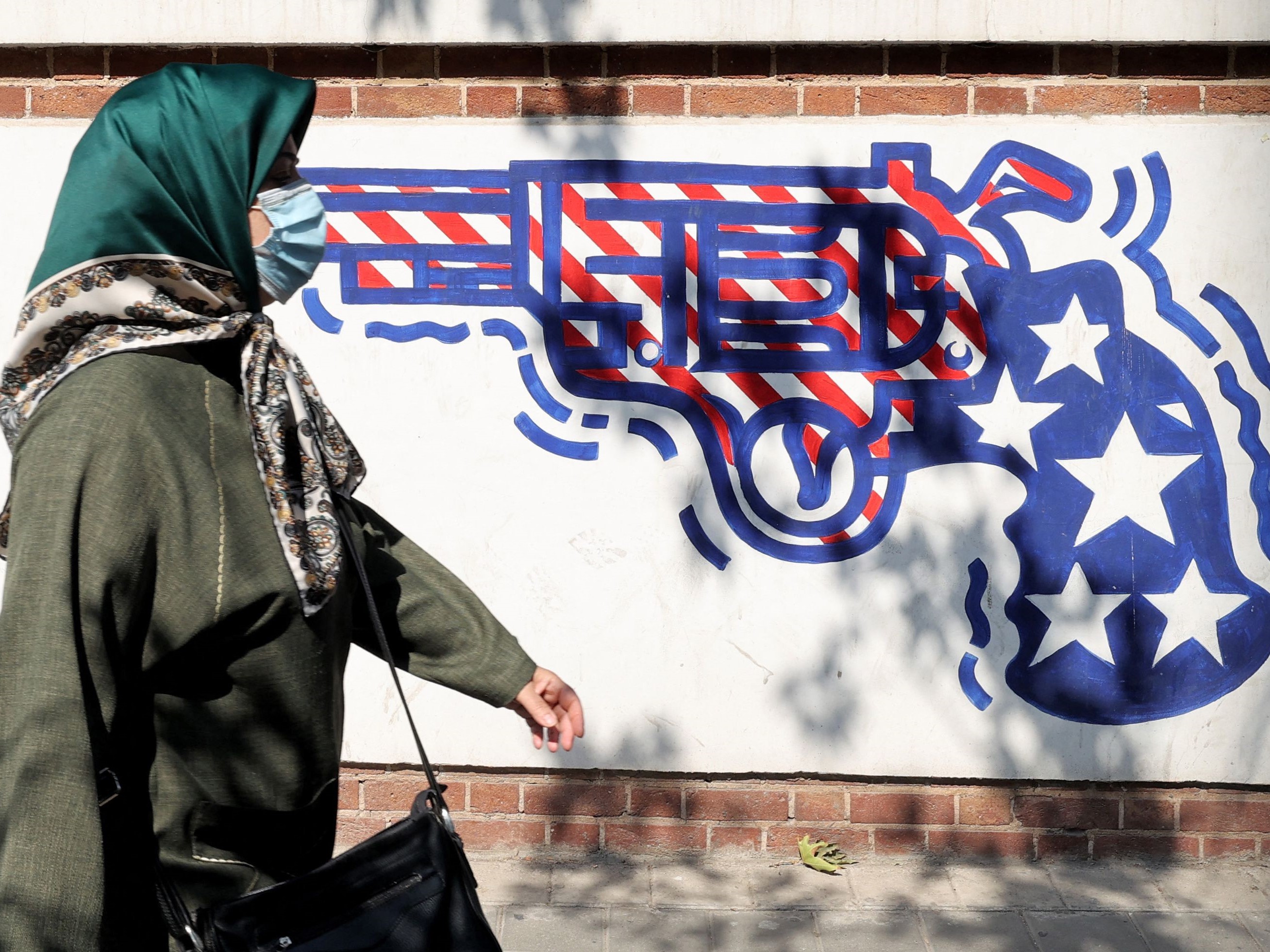Seorang wanita Iran bermasker berjalan melewati mural yang menggambarkan revolver yang dilukis dengan warna bendera AS, Teheran.