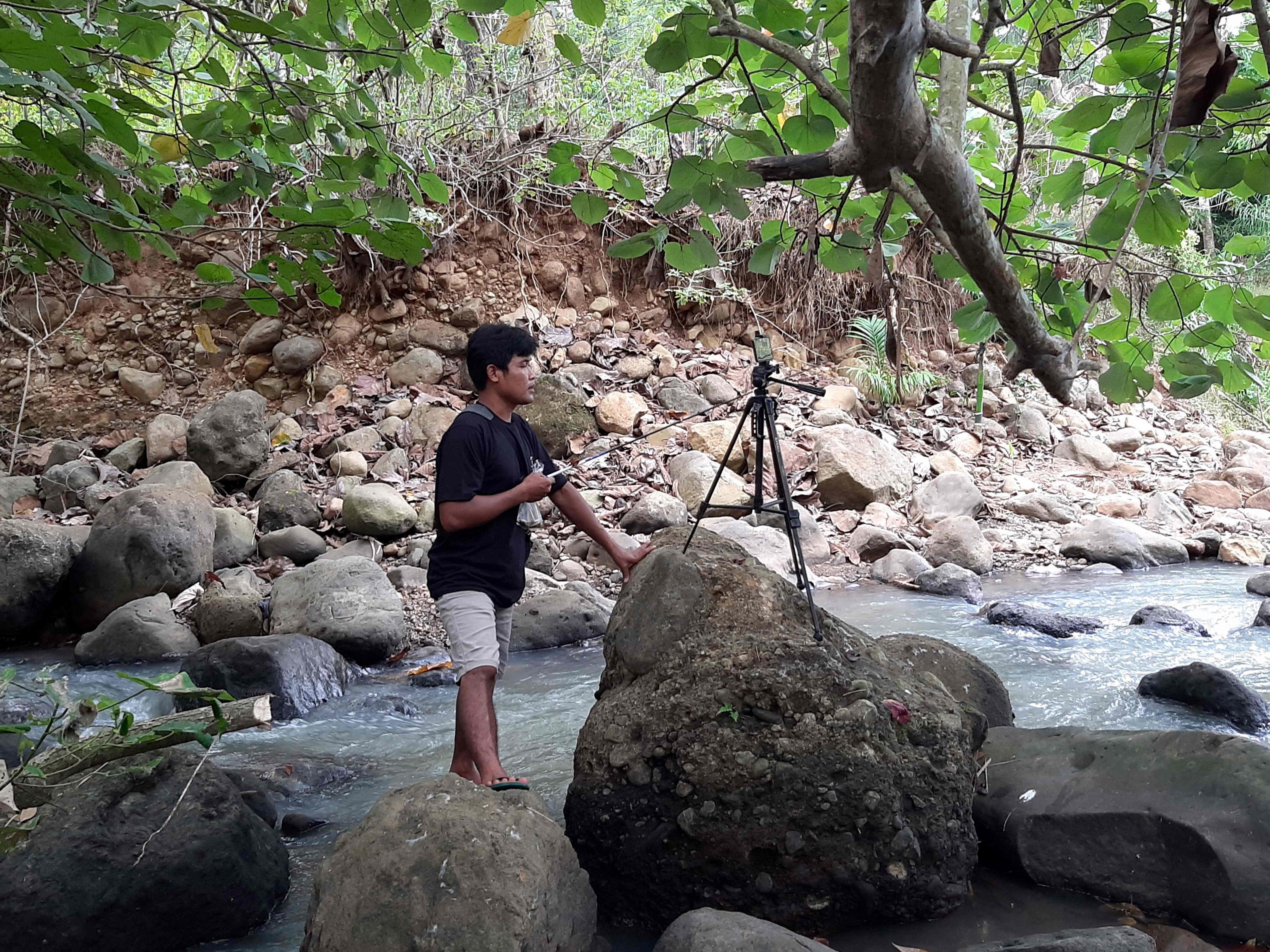 YouTuber Siswanto Indonesia merekam salah satu videonya di sebelah aliran air pada komunitas pertanian di Banyuwangi. 