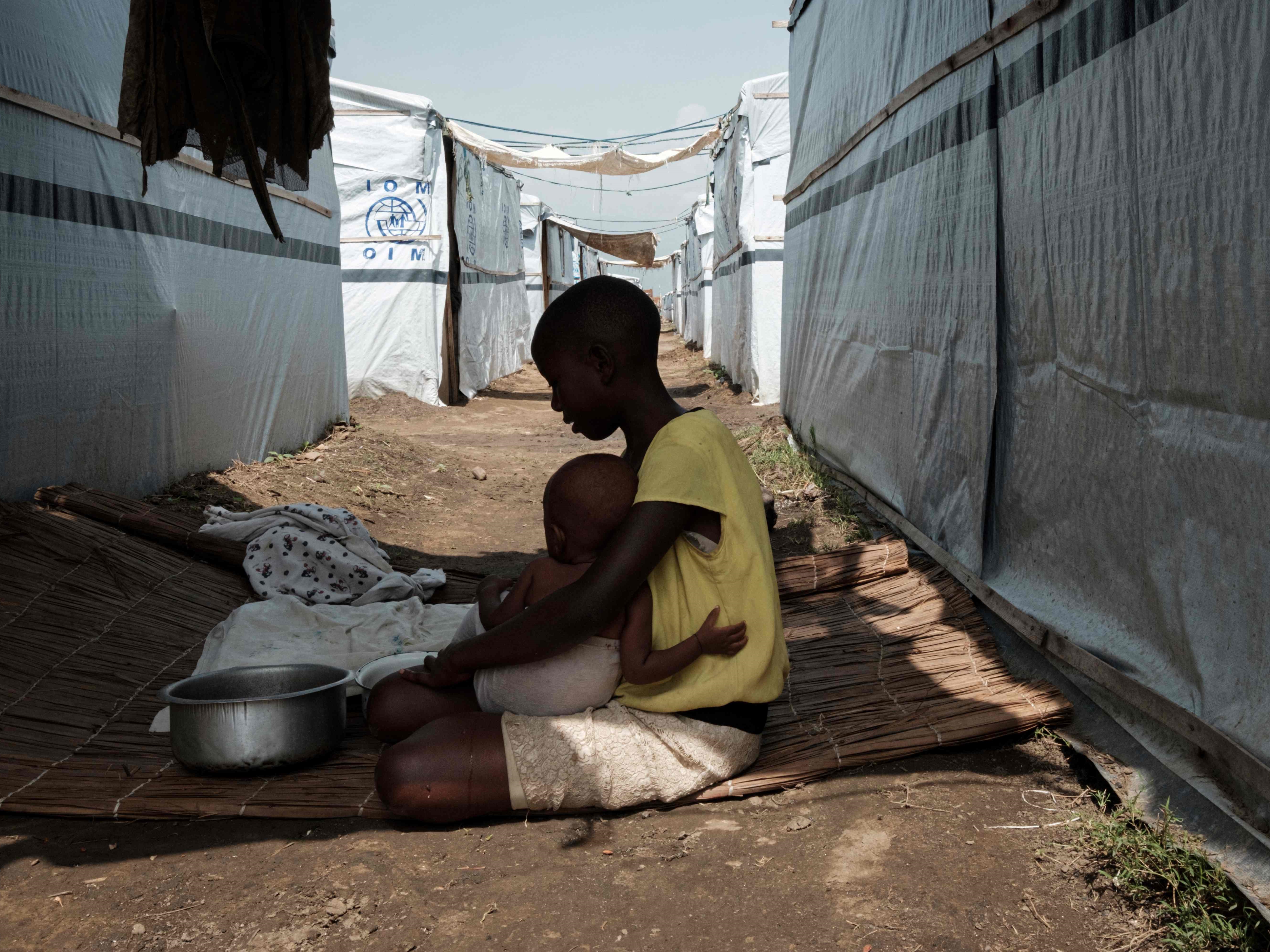 Seorang anak perempuan menggendong bayi dan bersiap memasak di bawah bayang-bayang tempat penampungan sementara di Burundi.