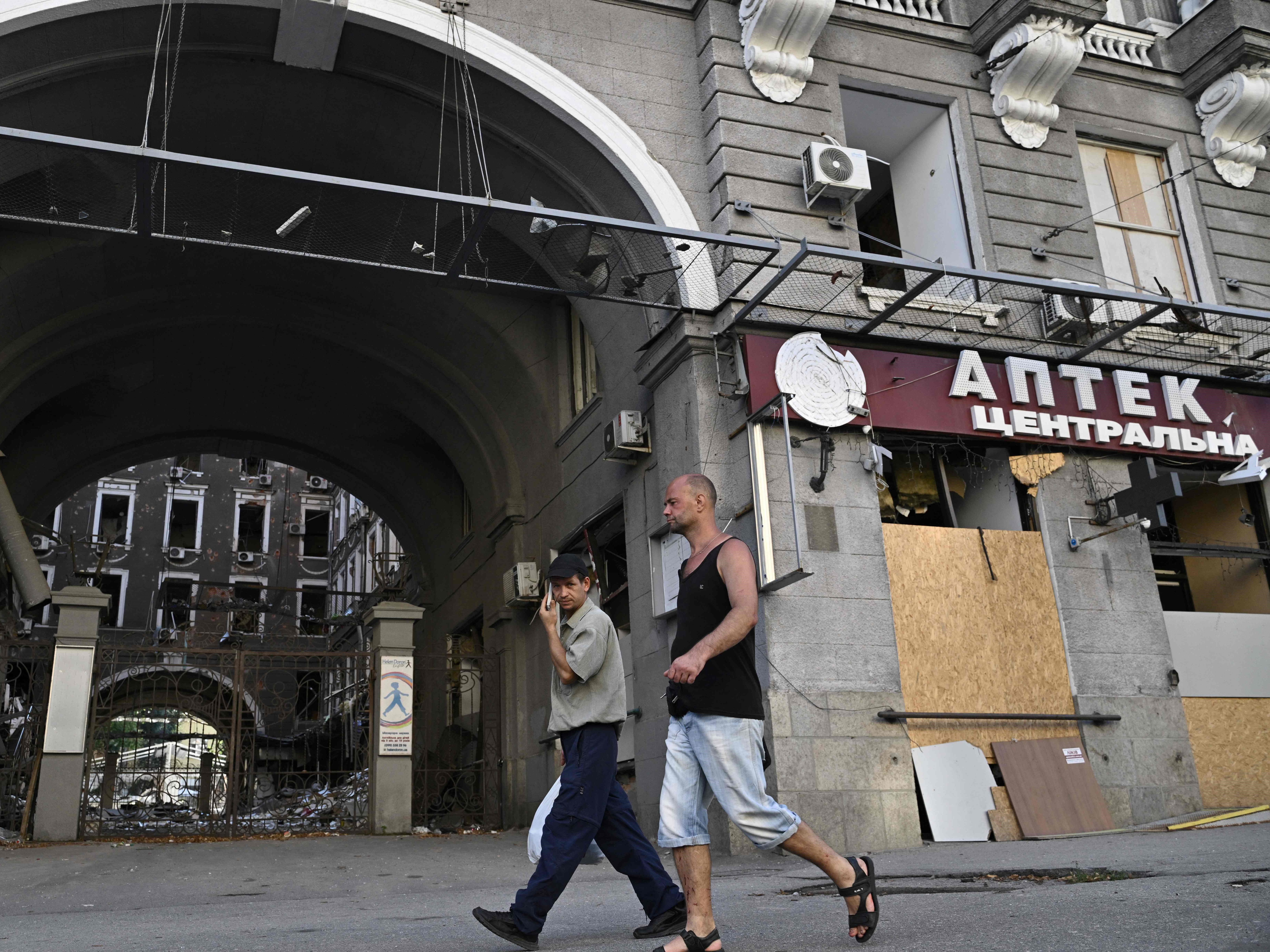 Dua pria berjalan melewati jendela tertutup toko obat yang tidak beroperasi setelah serangan di pusat Kharkiv, pada 27 Juli 2022.
