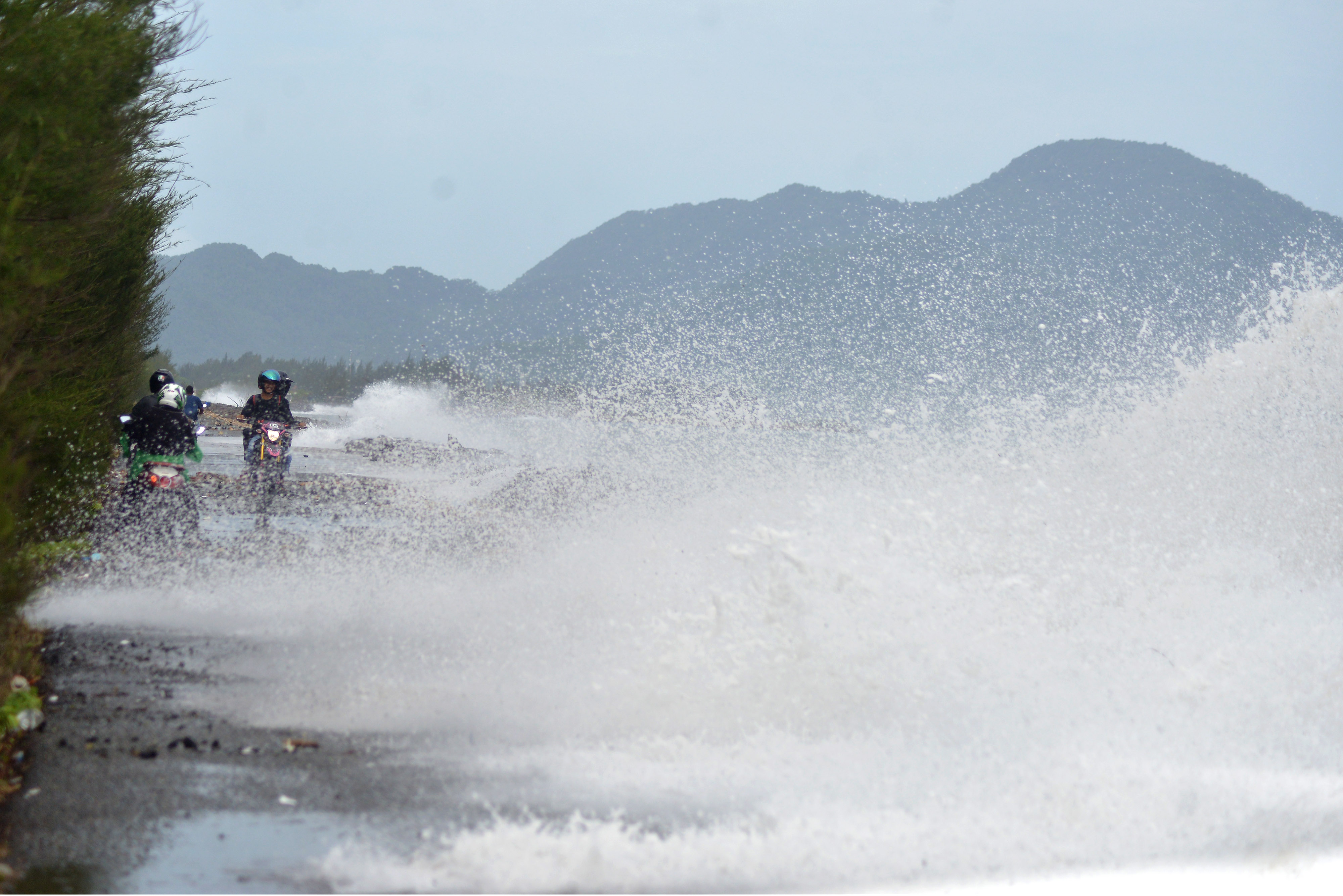 Pengendara motor melintasi jalan pantai saat berlangsung gelombang tinggi.