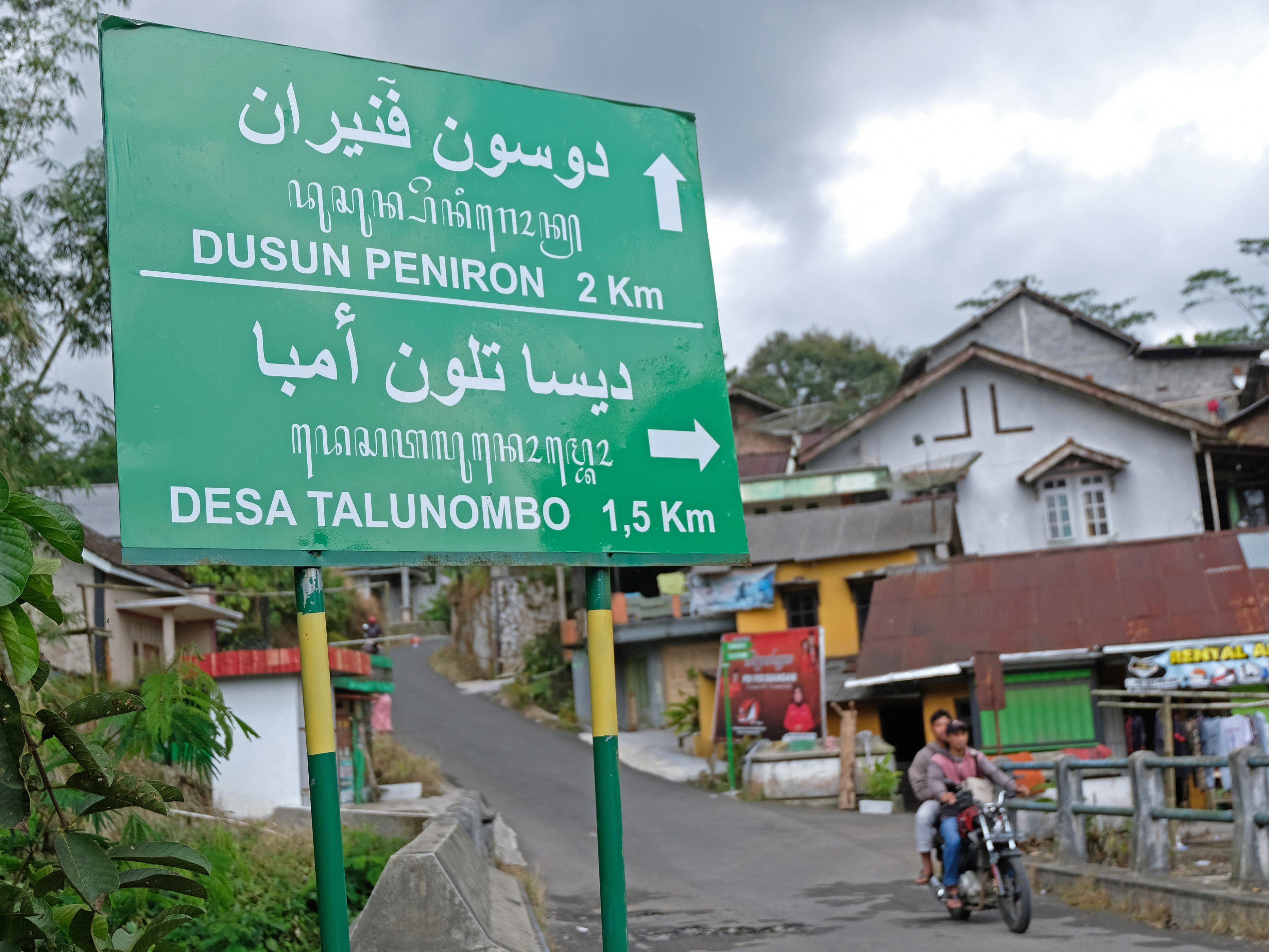 Warga melintasi papan penunjuk arah tiga bahasa di jalan Desa Talunombo, Sapuran, Wonosobo, Jawa Tengah, Sabtu (9/4/2022).