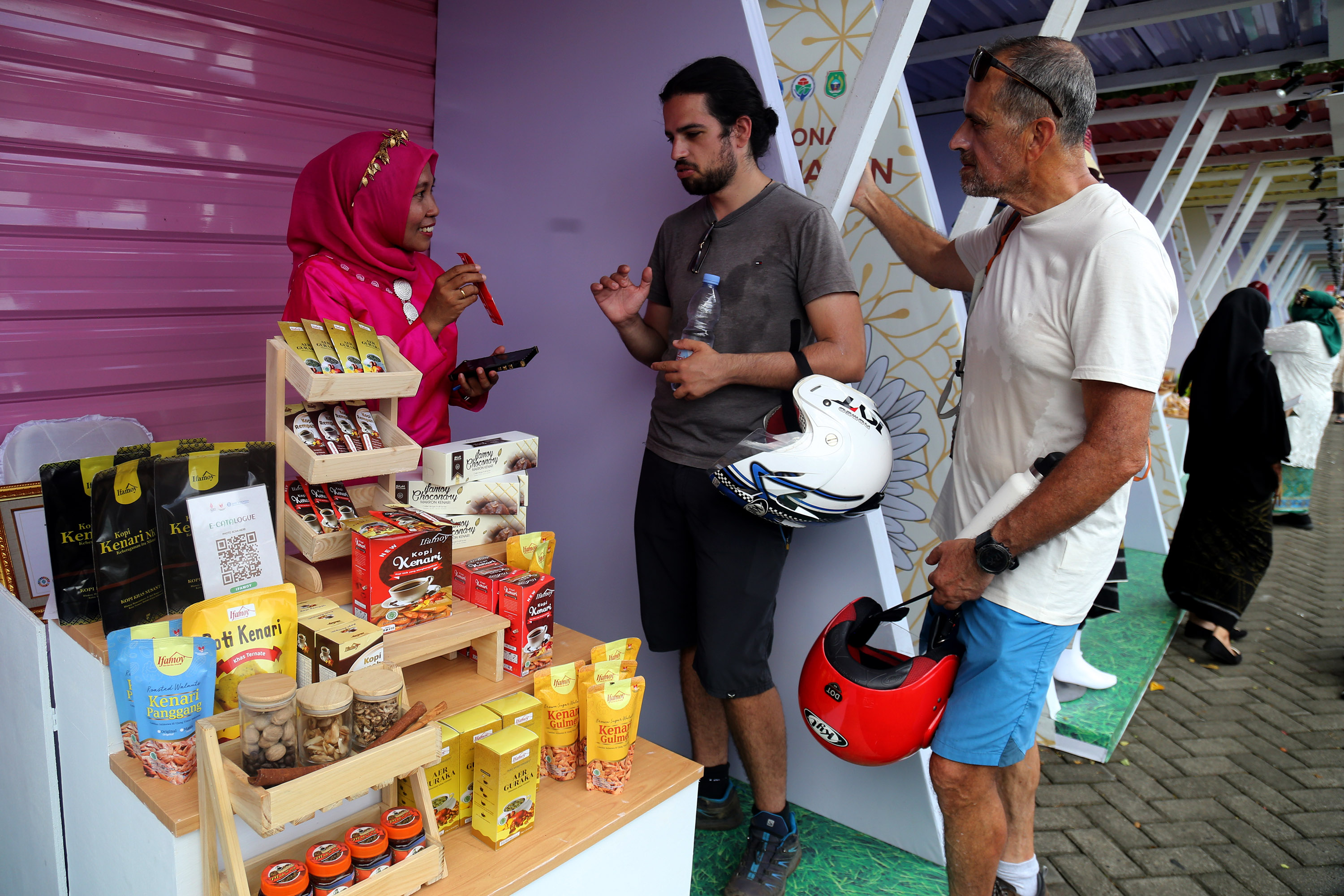 Penjaga stan menjelaskan salah satu produk UMKM kepada dua wisatawan asing.