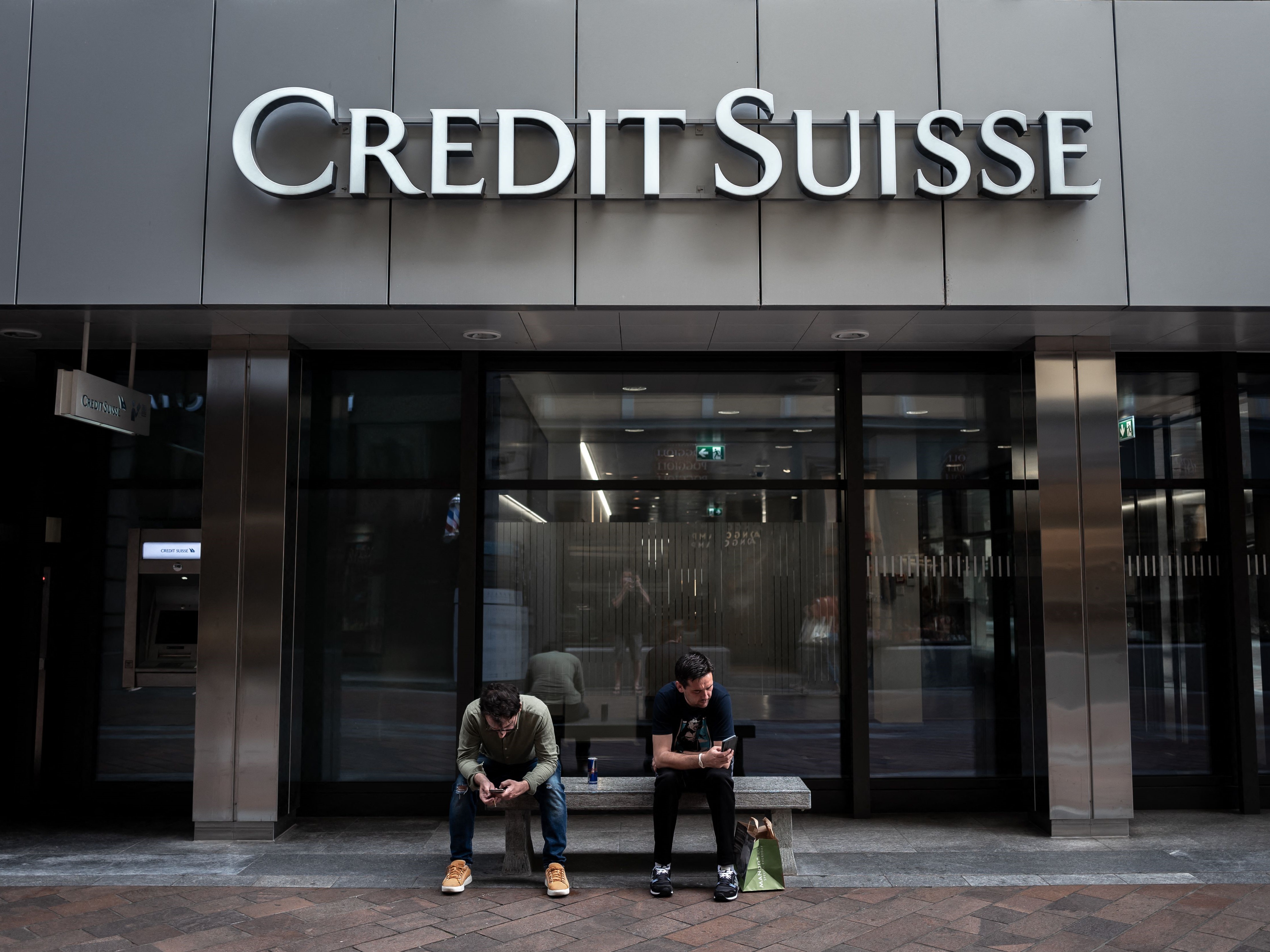 Dua pria duduk di bawah logo Credit Suisse yang dipajang suatu gedung di Lugano, Swiss, pada 9 Juni 2023.