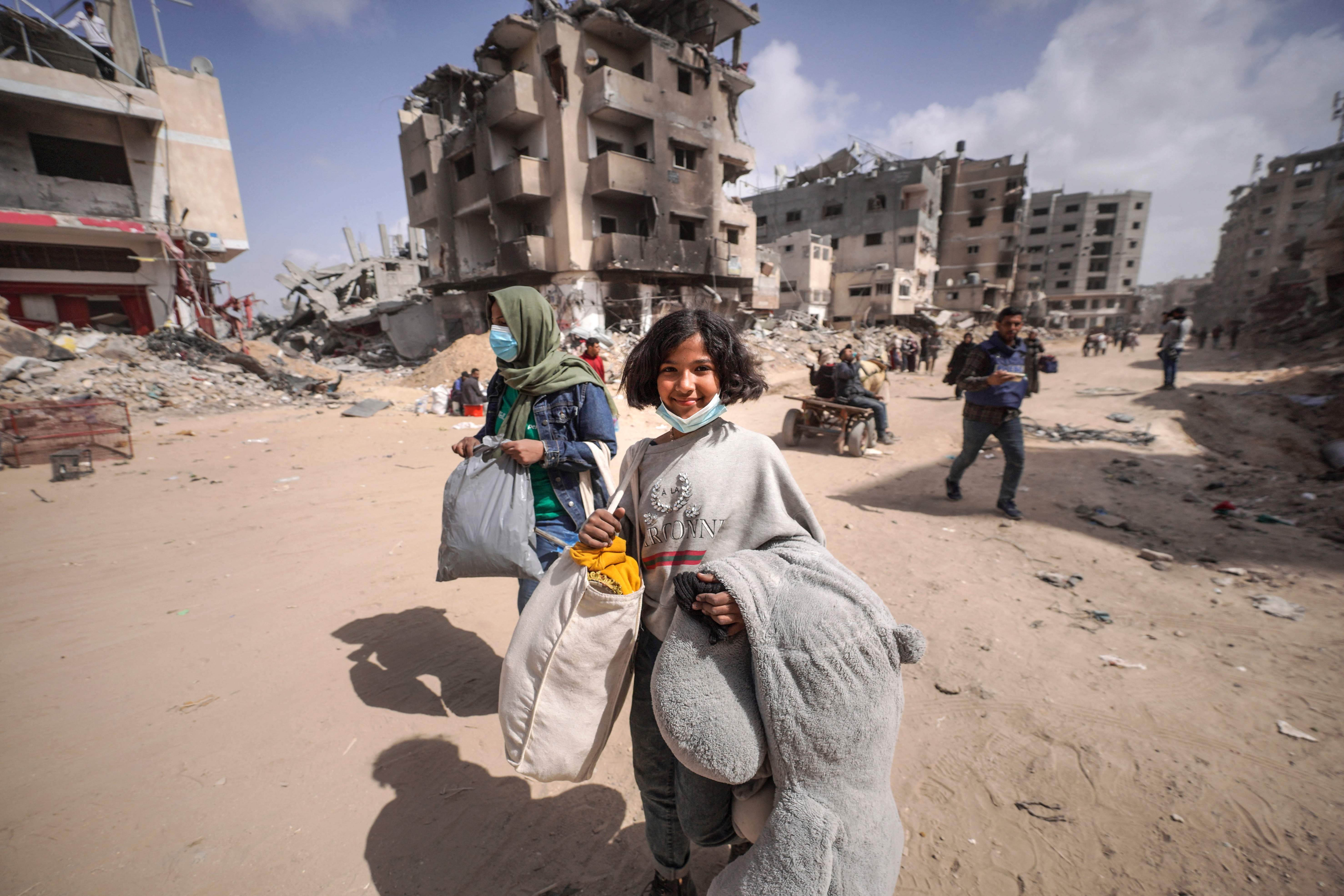 Pengungsi Palestina membawa barang-barang mereka melalui jalan dekat puing-puing bangunan yang hancur berkeping-keping