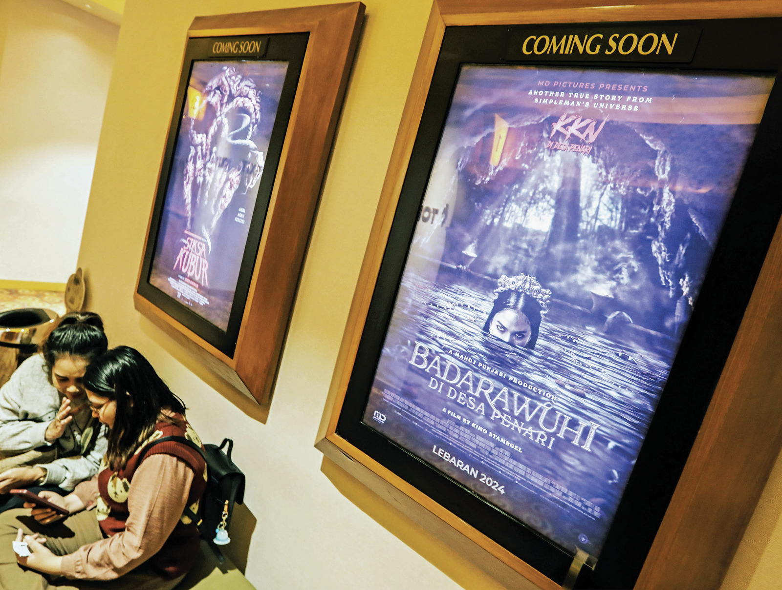 Pengunjung duduk menunggu jadwal tayangan film di bawah poster film horor Siksa Kubur dan Badarawuhi di Desa Penari yang terpasang di XXI.